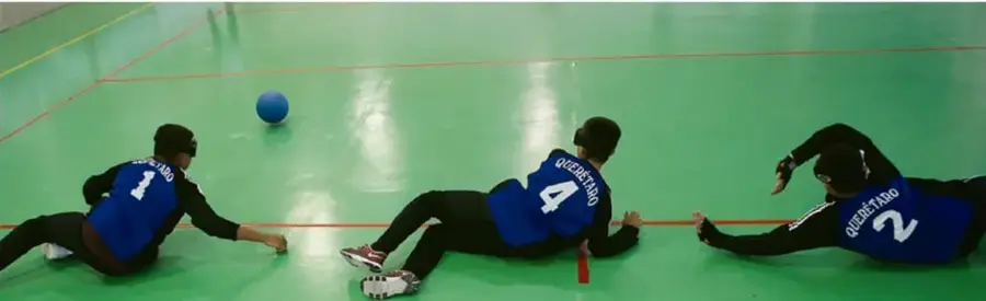 Fotografía de tres estudiantes en un patio de gimnasio con piso verde, vestidos con camisetas azules numeradas (1, 4 y 2) y antifaces negros, jugando sentados o arrastrándose en el suelo; hay una pelota azul en el piso.