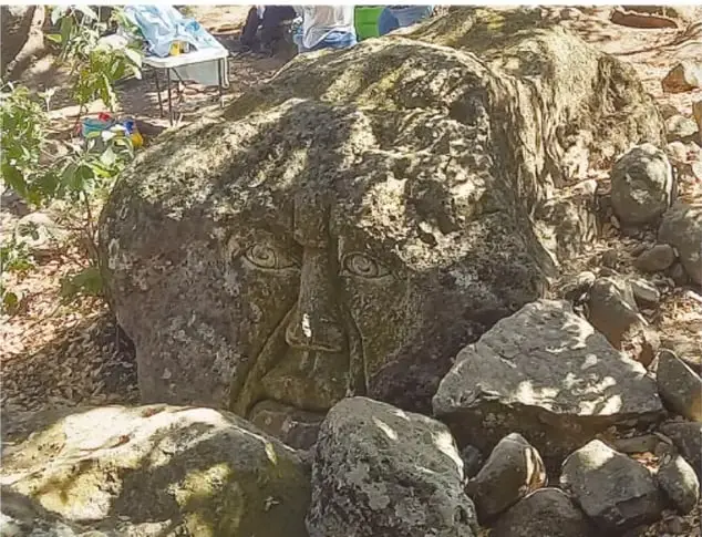 Fotografía de la segunda cabeza gigante (rostro de mujer) semioculta entre piedras cubiertas de musgo, en un área con árboles y objetos de visitantes al fondo.