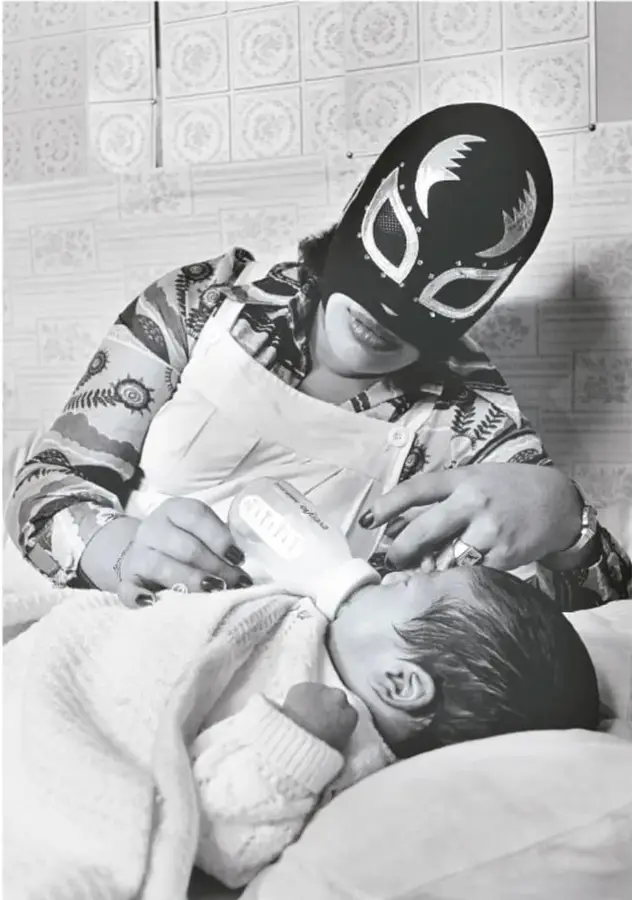 Fotografía en blanco y negro de una mujer con máscara de luchadora y delantal, dando un biberón a un bebé. Al pie dice: 'La Briosa y su hijo, 1984, Lourdes Grobet (1940-2022)'.