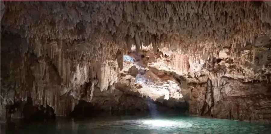 Fotografía del interior de una cueva con estalactitas y un cenote de agua turquesa iluminado.
