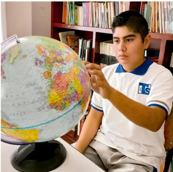 Fotografía de un estudiante de telesecundaria consultando un globo terráqueo frente a una estantería con libros; crédito: Luis Eduardo García Medel, Tlaxcala.