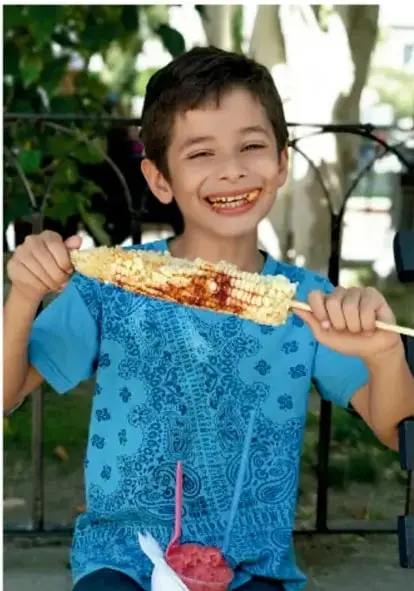 Niño sonriente sosteniendo un elote con salsa y limón, acompañado de un vaso con bebida roja.