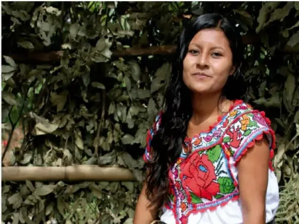 Fotografía de una joven indígena con blusa bordada de colores, frente a vegetación, que ilustra la diversidad cultural y la identidad.
