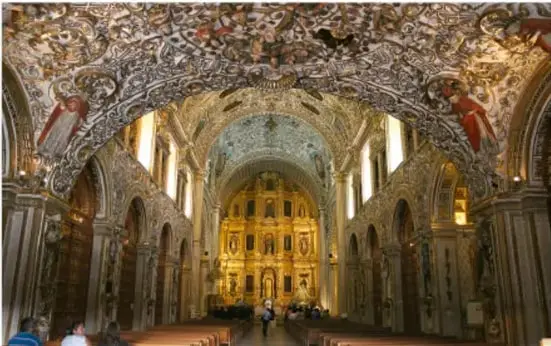 Interior de un templo virreinal con retablo dorado, bóvedas y arquitectura barroca lujosamente decorada.