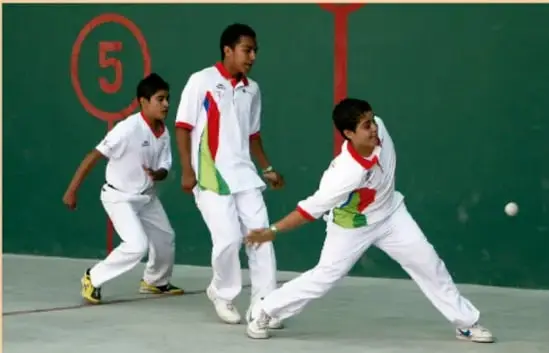 Fotografía de tres estudiantes en una cancha techada con pared verde (número 5 pintado): dos con uniforme blanco y uno con camiseta tricolor, practicando frontón con pelota en el aire.