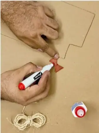 Fotografía de dos manos trabajando sobre cartón: una sostiene un marcador rojo y la otra recorta una figura con forma de flecha; alrededor hay un cordel enrollado y cinta adhesiva.