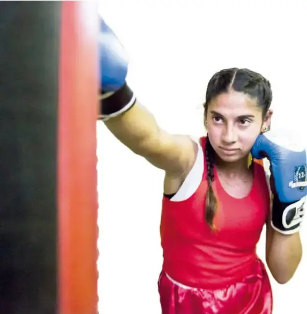Fotografía de una niña con guantes de boxeo azules y playera roja, con el brazo extendido hacia un costal de entrenamiento.