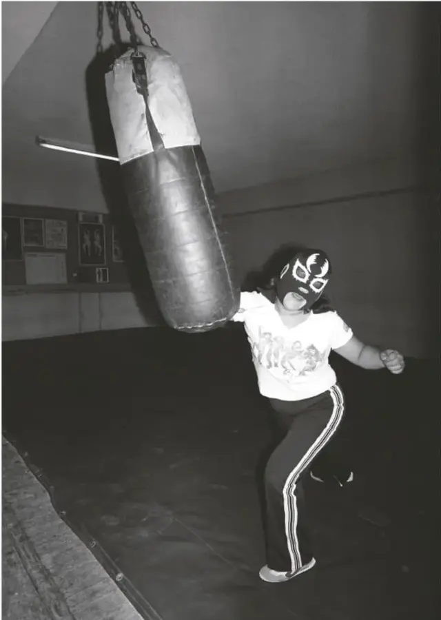 Fotografía en blanco y negro de una luchadora enmascarada golpeando un costal colgado del techo con cadenas. Viste playera blanca con estampado y pants oscuro con franjas laterales. El fondo muestra un muro con carteles y un piso oscuro de gimnasio. Pie de foto: 'La Briosa entrenando. 1982. Lourdes Grobet (1940-2022)'.