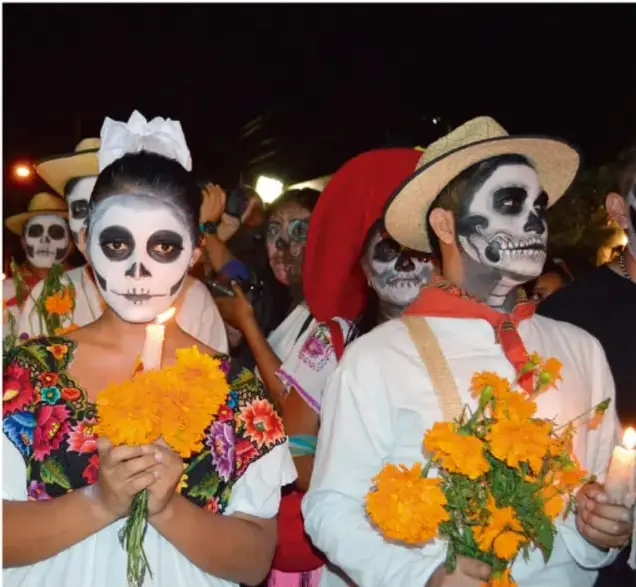 Fotografía nocturna de un grupo de personas con el rostro pintado como calaveras, portando ramos de flores de cempasúchil naranjas y velas encendidas, en un festejo tradicional mexicano (probablemente Día de Muertos).