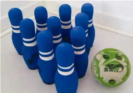 Fotografía de bolos azules de boliche con franjas blancas y una pelota verde con vetas negras, junto a una pared blanca.