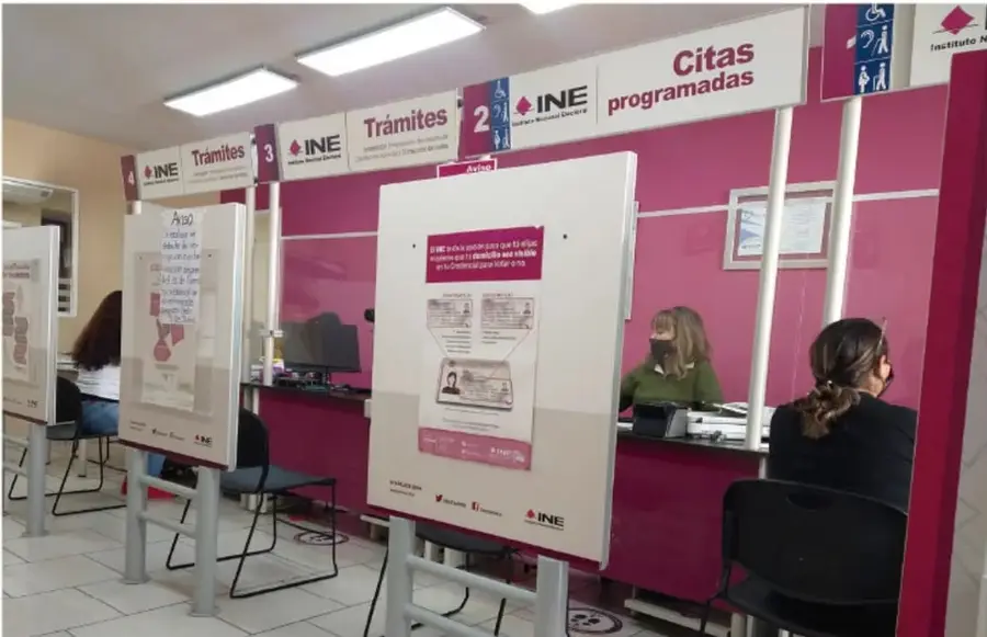 Fotografía del interior de una oficina del INE (Instituto Nacional Electoral) en México: módulos de atención con mamparas magenta y carteles 'Trámites' numerados y 'Citas programadas'; una empleada con cubrebocas atiende a una ciudadana en el mostrador; en los mamparos hay infografías sobre el trámite de credencial para votar.