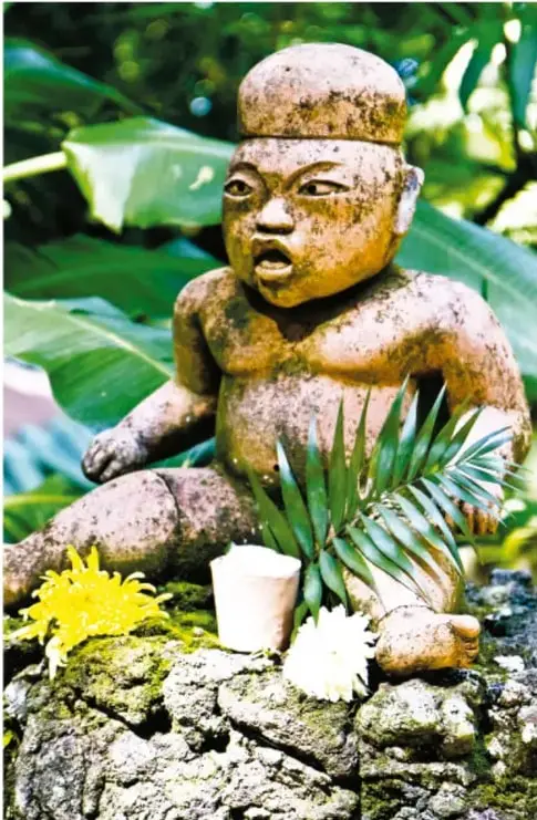 Escultura de piedra prehispánica (cabeza y torso de figura antigua) entre plantas verdes, adornada con flores amarillas y blancas y una taza con veladora.