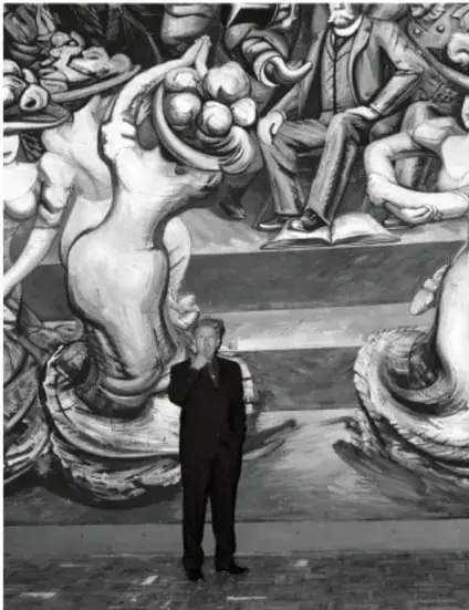Fotografía en blanco y negro de un hombre de traje frente a un mural con figuras humanas y elementos simbólicos.