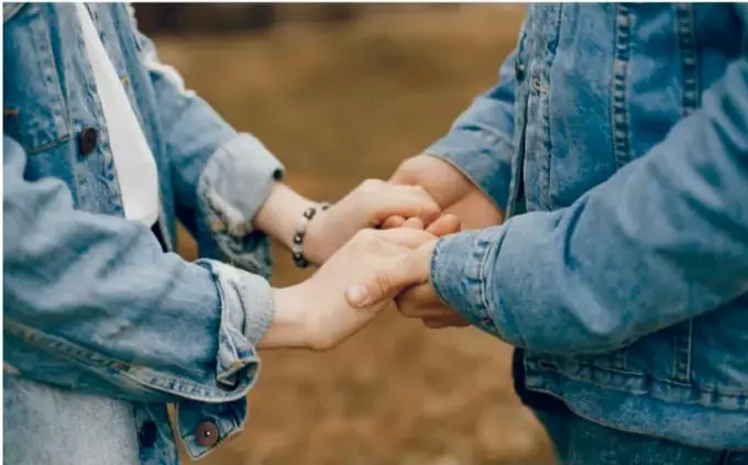 Fotografía de dos personas (aparentemente adolescentes o adultos jóvenes con chaquetas de mezclilla) tomadas de las manos en gesto de apoyo, acompañamiento y confianza.