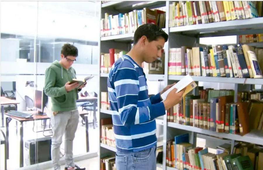 Fotografía de dos estudiantes leyendo libros de pie entre estanterías de una biblioteca escolar, uno con sudadera verde y otro con camisa azul a rayas.