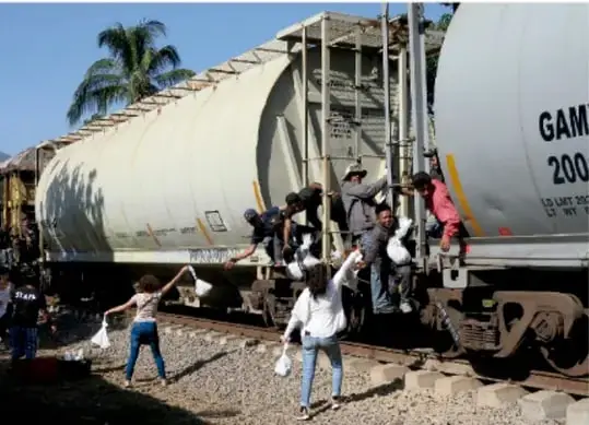 Fotografía de personas subidas y colgadas de un tren de carga (vagón tanque), en un entorno rural con palmeras; representa a migrantes que viajan sobre el tren, fenómeno conocido como 'la bestia' en la ruta migratoria hacia Estados Unidos.