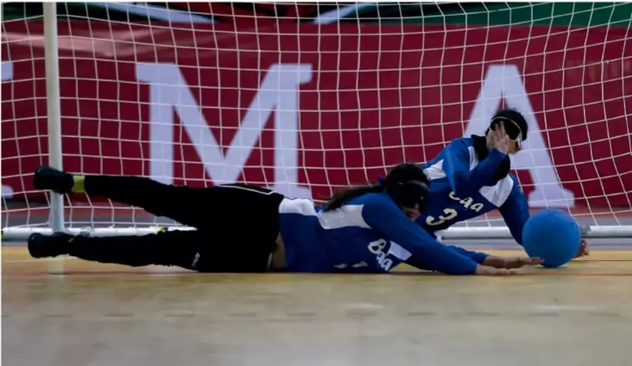 Fotografía de dos jugadoras con antifaz (deporte de colaboración-confrontación, tipo goalball) tumbadas en el piso alcanzando una pelota azul junto a la portería con red.