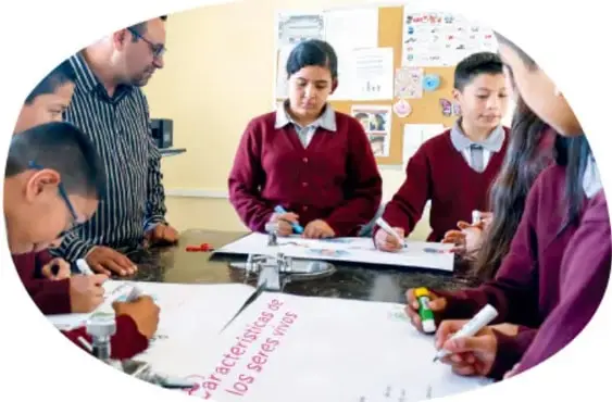 Fotografía circular de un grupo de estudiantes en uniforme y su docente trabajando en equipo alrededor de una mesa, escribiendo y coloreando en hojas; un cartel muestra el texto '...argentísimos los sueños'.