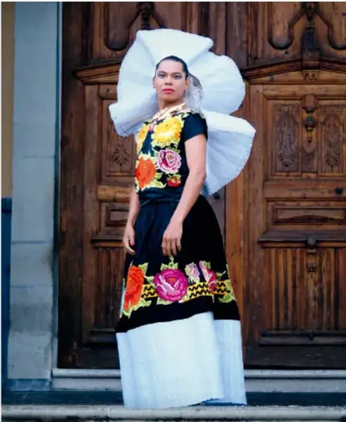 Persona con vestimenta tradicional del Istmo de Tehuantepec, con falda bordada y tocado blanco, frente a una puerta de madera.