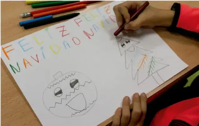 Fotografía de las manos de un estudiante que colorea con lápices de colores una hoja con el texto «FELIZ NAVIDAD» y dibujos de un árbol navideño y un muñeco de nieve.