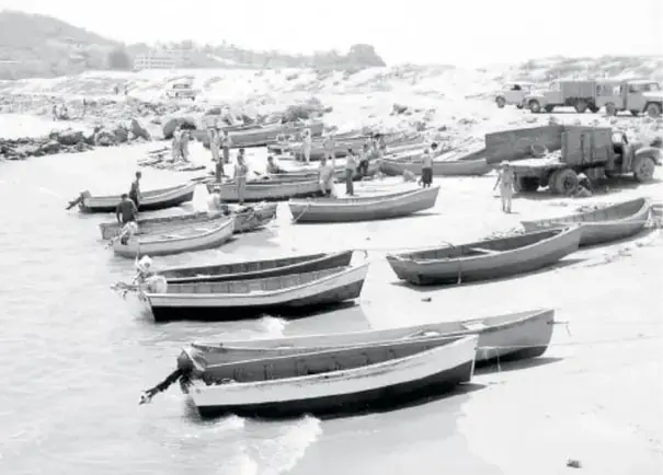 Fotografía en blanco y negro de muchos botes o barcas pequeñas de madera varadas en una playa, con camiones al fondo; evoca los transportes del pasado.
