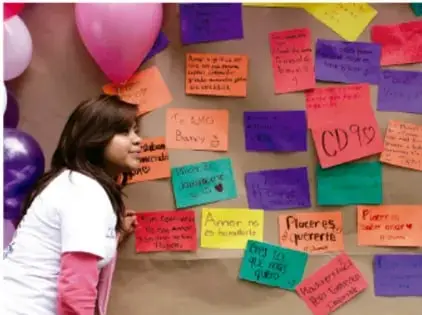 Fotografía de una estudiante frente a un muro de la comunidad lleno de notas de colores con mensajes contra la violencia y por la paz, con globos de colores alrededor.