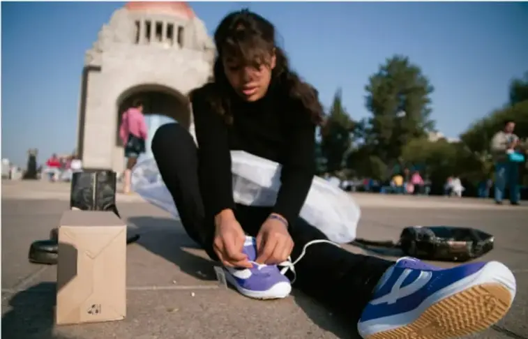 Fotografía de una joven sentada en el piso en un espacio público (monumento al fondo, Monumento a la Revolución en México), amarrándose los zapatos deportivos, preparándose para correr o hacer ejercicio al aire libre.
