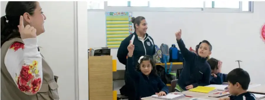Fotografía de una maestra y estudiantes en un aula, uno de ellos levanta la mano.