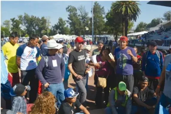 Fotografía de un grupo numeroso de personas migrantes caminando al aire libre, con mochilas y gorras, bajo un cielo despejado.