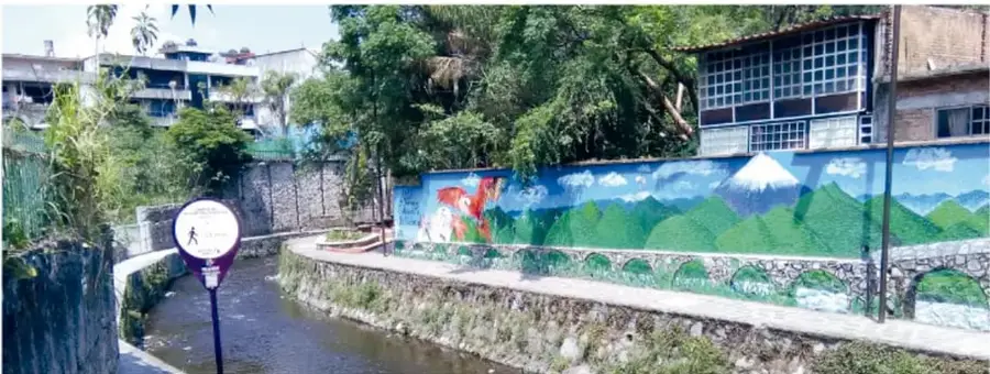 Fotografía de un muro pintado junto a un río urbano con montañas verdes, un volcán con nieve y nubes; hay un letrero turístico redondo al borde del agua.