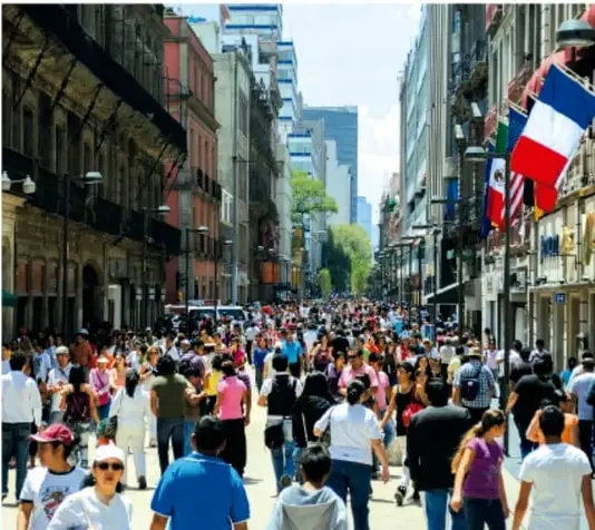 Fotografía de una calle muy concurrida de una ciudad mexicana (posiblemente el centro histórico de la Ciudad de México) con multitud de peatones caminando y banderas colgando de los edificios, ilustrando el tema 'De la multitud a la comunidad'.