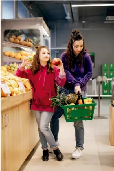 Fotografía de una madre y su hija comprando frutas y verduras en el mercado; la niña sostiene una manzana y la madre una canasta verde con piña y otras frutas.