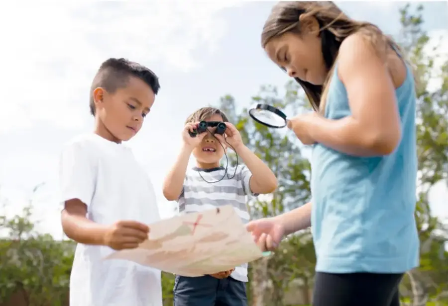Fotografía de tres niños al aire libre: dos observan un mapa de papel (uno con binoculares) y una niña lo examina con una lupa.
