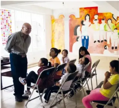 Fotografía de un grupo de personas sentadas en círculo dentro de un aula, dialogando con un adulto mayor de pie.
