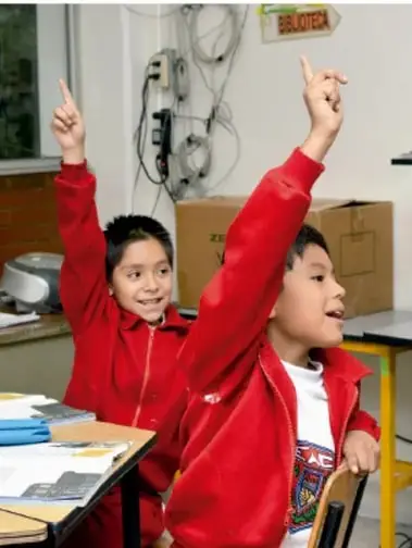 Fotografía de dos niños con uniforme rojo levantando la mano en un salón de clases.