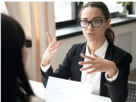 Fotografía de una mujer con lentes y saco negro que conversa con otra persona, posiblemente en una entrevista de trabajo o reunión.