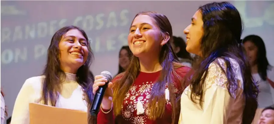 Fotografía de tres jóvenes sonrientes en un evento comunitario o escolar; una sostiene un micrófono y otra una carpeta; al fondo se lee parcialmente 'GRANDES COSAS...'. Ilustra la participación comunitaria.