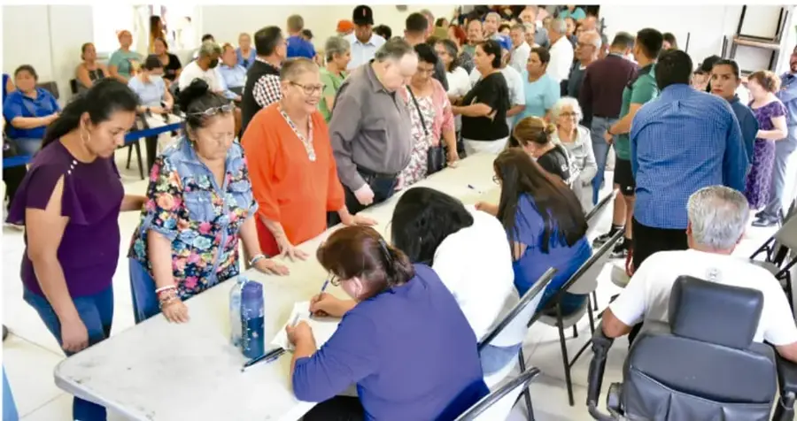Fotografía de una asamblea comunitaria: ciudadanos de distintas edades esperan en mesas donde personal registra su participación.
