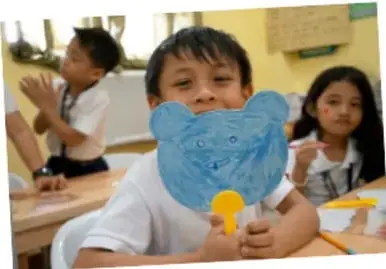 Fotografía de estudiantes de primaria en el aula; un niño sostiene una máscara de papel azul con forma de animal y una niña observa sonriente.