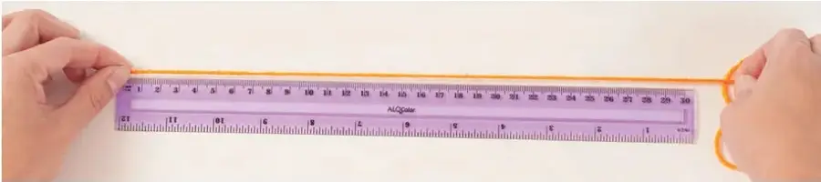 Dos manos sostienen una regla plástica morada de 30 cm; un trozo de lana naranja se extiende a lo largo de la regla desde el extremo izquierdo hasta cerca de la marca de 29-30 cm.