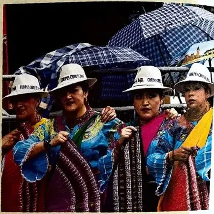 Fotografía de varias mujeres indígenas con vestimenta tradicional, sombreros y paraguas, en un espacio público.