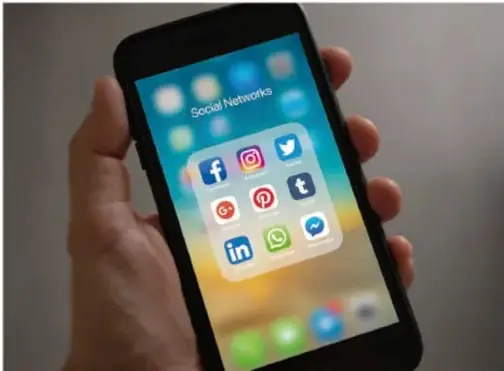 Fotografía de una mano sosteniendo un teléfono inteligente que muestra una carpeta llamada 'Social Networks' con íconos de Facebook, Instagram, Twitter, Pinterest, Tumblr, LinkedIn y WhatsApp.