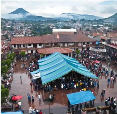 Fotografía de una plaza pública con un tianguis o mercado al aire libre, con personas caminando y puestos con lonas azules. Al fondo se observan montañas y edificios.