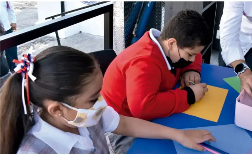 Fotografía de estudiantes de primaria trabajando en una mesa con hojas de colores; uno escribe y otra observa. Usan cubrebocas.