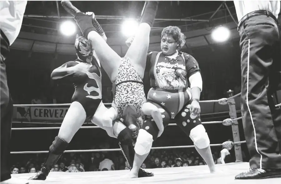 Fotografía en blanco y negro de una lucha libre femenil: tres luchadoras sobre el cuadrilátero, una de ellas levantada por las piernas; al fondo el público, las cuerdas y reflectores; a los lados, piernas de personas de pie.