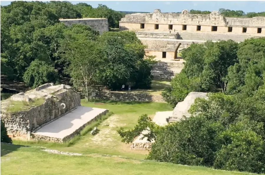 Fotografía panorámica de una zona arqueológica: un patio hundido con estructuras de piedra a los lados (aparente juego de pelota) rodeado de árboles y edificios prehispánicos al fondo.