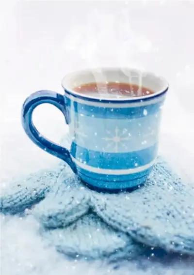 Fotografía de una taza de té humeante sobre un suéter azul con copos de nieve, en ambiente invernal.