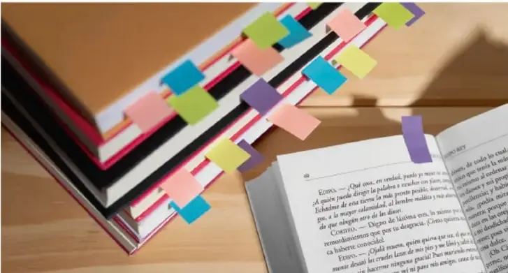 Fotografía de libros con cintas de colores (marcapáginas) sobre un escritorio junto a un libro abierto, aludiendo a la lectura y el estudio de textos.