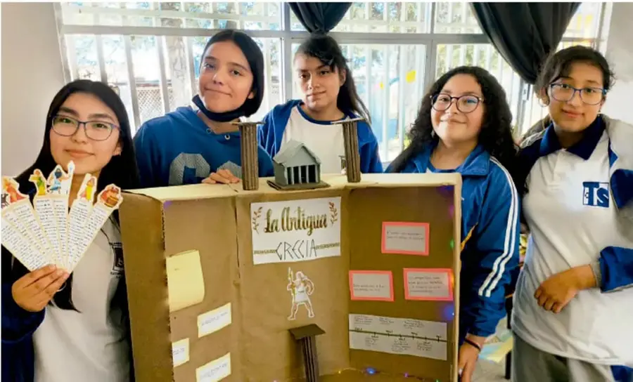Fotografía de cinco estudiantes de telesecundaria de uniforme, sonriendo, junto a una maqueta-caja de cartón del proyecto "La Antigua Grecia", con fichas y una línea del tiempo impresas. Crédito: Luis Eduardo García Medel, Tlaxcala.