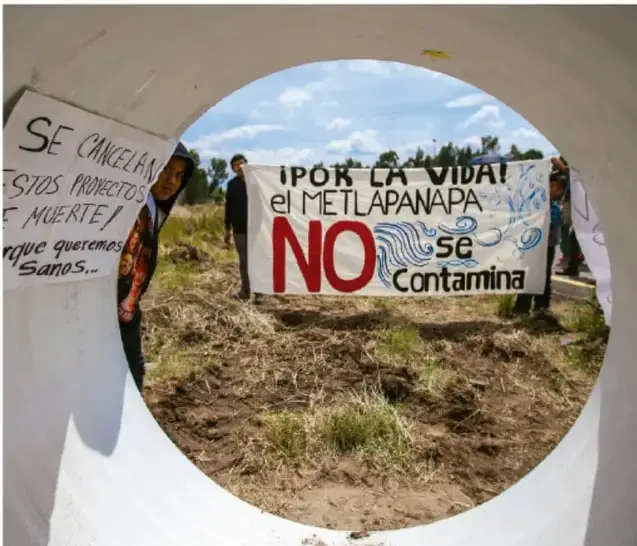 Fotografía de una protesta comunitaria: manifestantes sostienen carteles que dicen "¡Por la vida! el Metlapanapa NO se contamina" y "Se cancelan estos proyectos, ¿qué queremos, sanos... muerte!?". La imagen está enmarcada por un círculo blanco tipo mirilla.